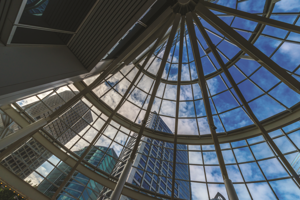 The image shows a view from inside a building looking up through a glass dome ceiling. The glass panels form a geometric pattern, revealing a cityscape of modern skyscrapers and a partly cloudy blue sky. The architecture captures the blend of urban design and natural light.