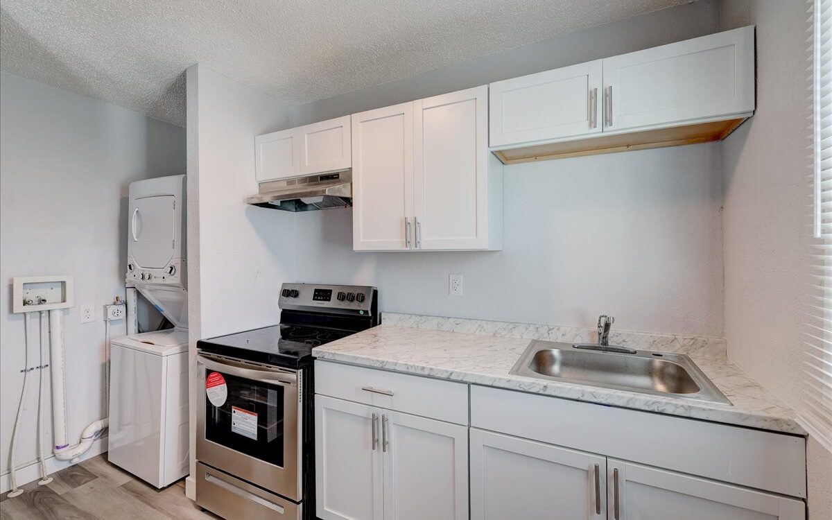 A compact kitchen features white cabinets, a stainless steel sink, and a marble-like countertop. A silver oven with a range hood is present. To the left, a stacked washer and dryer unit is installed. The space is brightly lit by natural light from an unseen source.