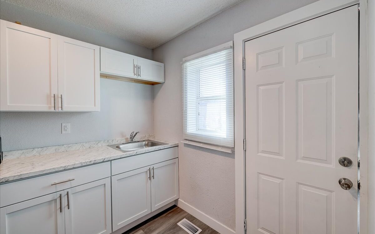 A small, bright kitchen with white cabinets and countertops. A stainless steel sink is situated under a window with closed blinds. There's a door with a window to the right, and the floor is finished with wood-like vinyl planks.