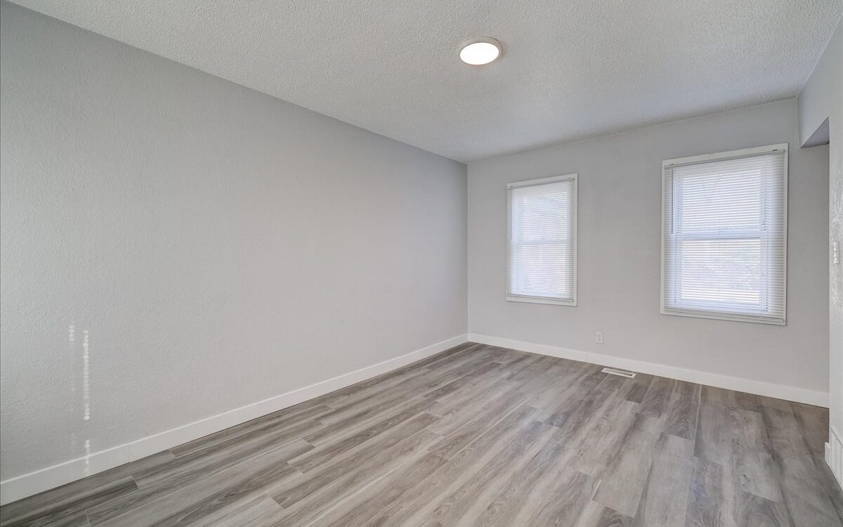 A minimalist, unfurnished room with light grey walls and two windows covered by blinds. The room features a ceiling light and grey hardwood flooring. Sunlight streams in, casting subtle shadows on the floor.