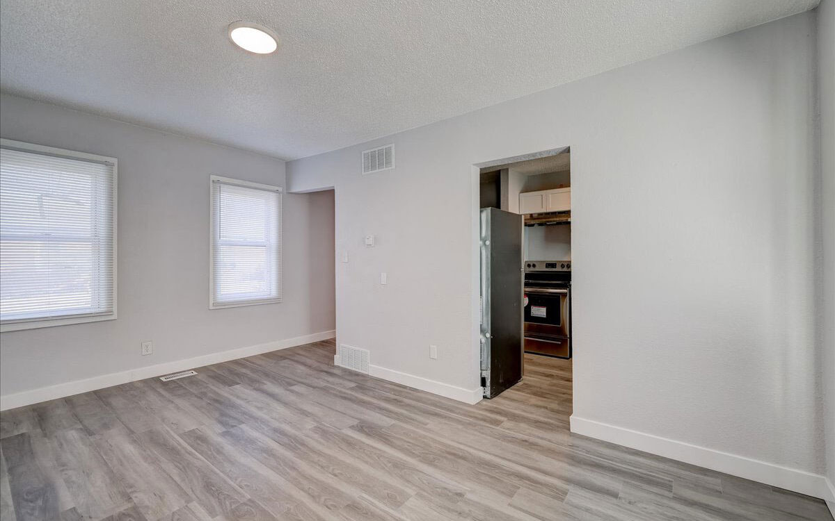 A modern, empty room with light gray walls and wood flooring. Two windows with white blinds on the left wall allow natural light. An open doorway in the middle right leads to a kitchen with stainless steel appliances. A round ceiling light is visible.