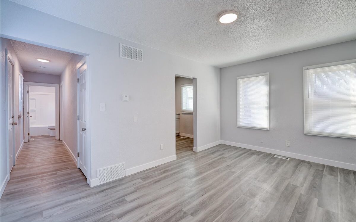 A living area with light gray walls, large windows, and light wood flooring. There is a hallway to the left leading to other rooms and a partially visible kitchen through a doorway. Recessed lighting and natural light brighten the space.