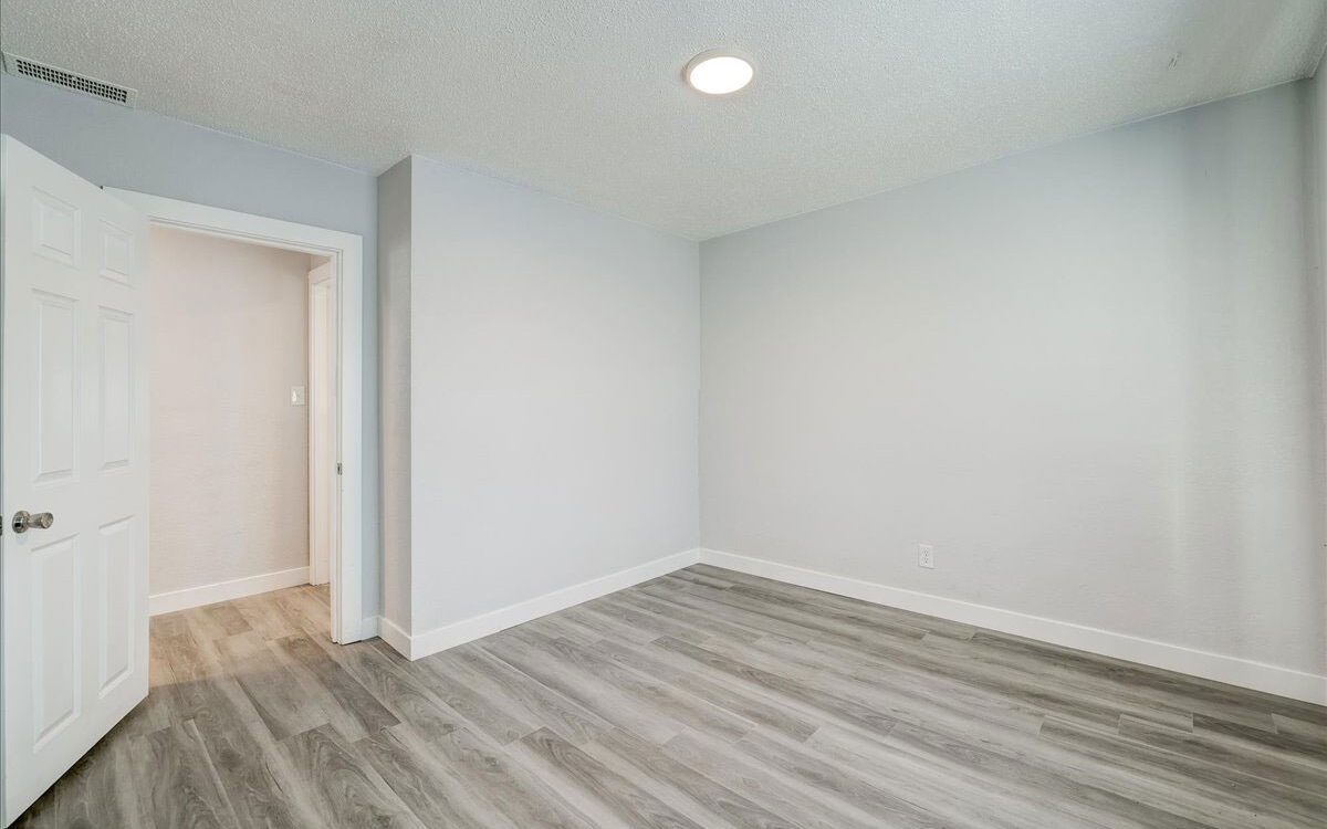 A minimalistic empty room with light-colored walls and a single overhead light. The floor is covered in gray-toned wood laminate. A partially open door leads to another space, with a glimpse of a hallway in the background.