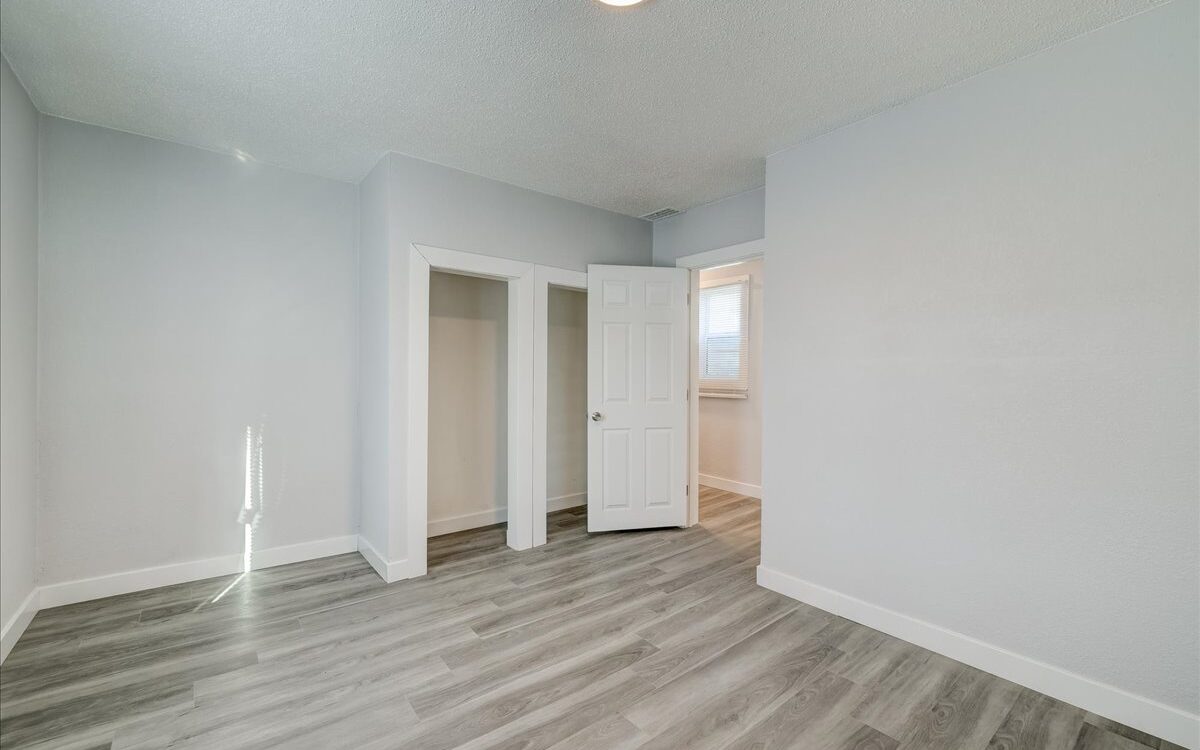 A small, empty room with light gray walls and wood-like vinyl flooring. It features a single door with a small closet on one side and an adjacent hallway. Sunlight streams in through an unseen window, casting light onto the left wall.