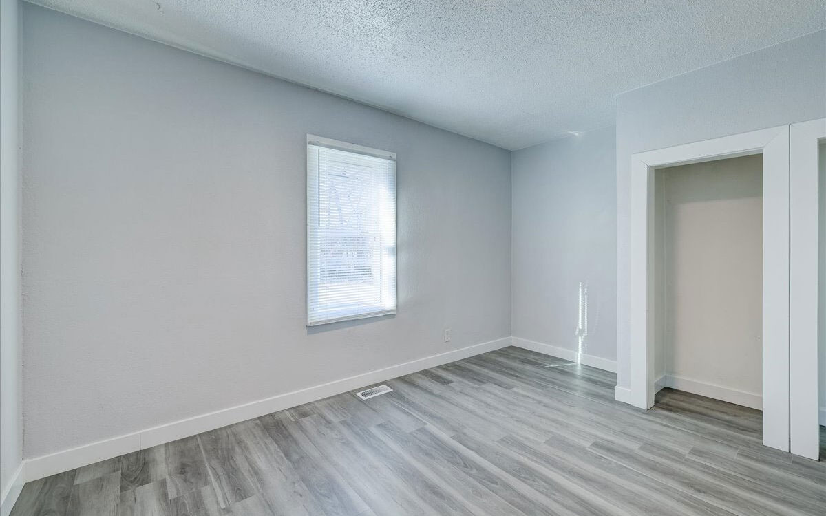 A small, empty room with light gray walls and a white-trimmed window letting in natural light. The floor is covered with gray wood-look vinyl planks. There’s an open closet space on the right and a ceiling vent visible near the window.