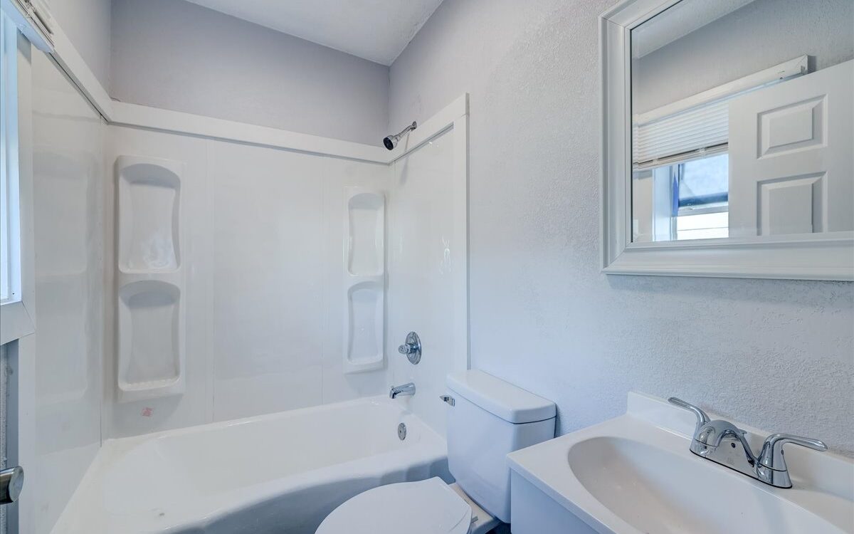 A modern bathroom features a white bathtub-shower combo with built-in shelving, a white toilet, and a white sink with a chrome faucet. Above the sink is a rectangular mirror with a white frame. The walls are light gray, and the room is well-lit with natural light.