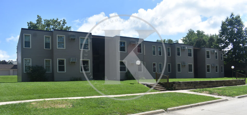 A two-story apartment building with a grey exterior and multiple windows. The building is set in a grassy area with a sidewalk in the foreground. There are trees and clouds in the background, reflecting a clear day.