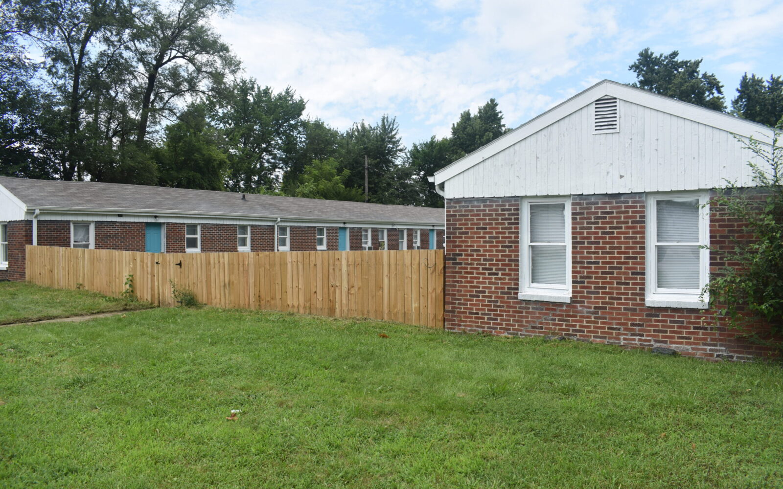 A grassy lawn in front of a brick building with white trim and two windows. A wooden fence separates the front building from several similar brick buildings in the background. Tall trees are visible beyond the buildings under a partly cloudy sky.