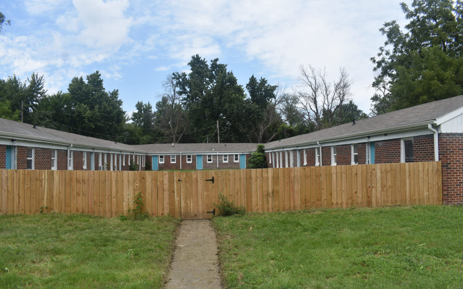 A courtyard is surrounded by one-story brick buildings with white trim. The courtyard has a path leading to a wooden fence gate. The area is grassy and bordered by trees. The sky above is partly cloudy.