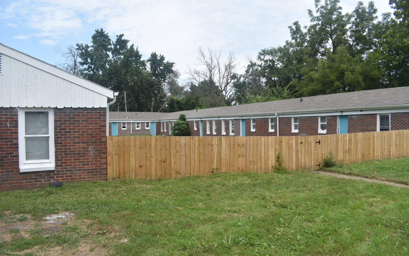 A grassy yard is bordered by a wooden fence, behind which are single-story, red brick buildings with white trim and gray roofs. Trees and a partly cloudy sky are in the background, suggesting a residential area.