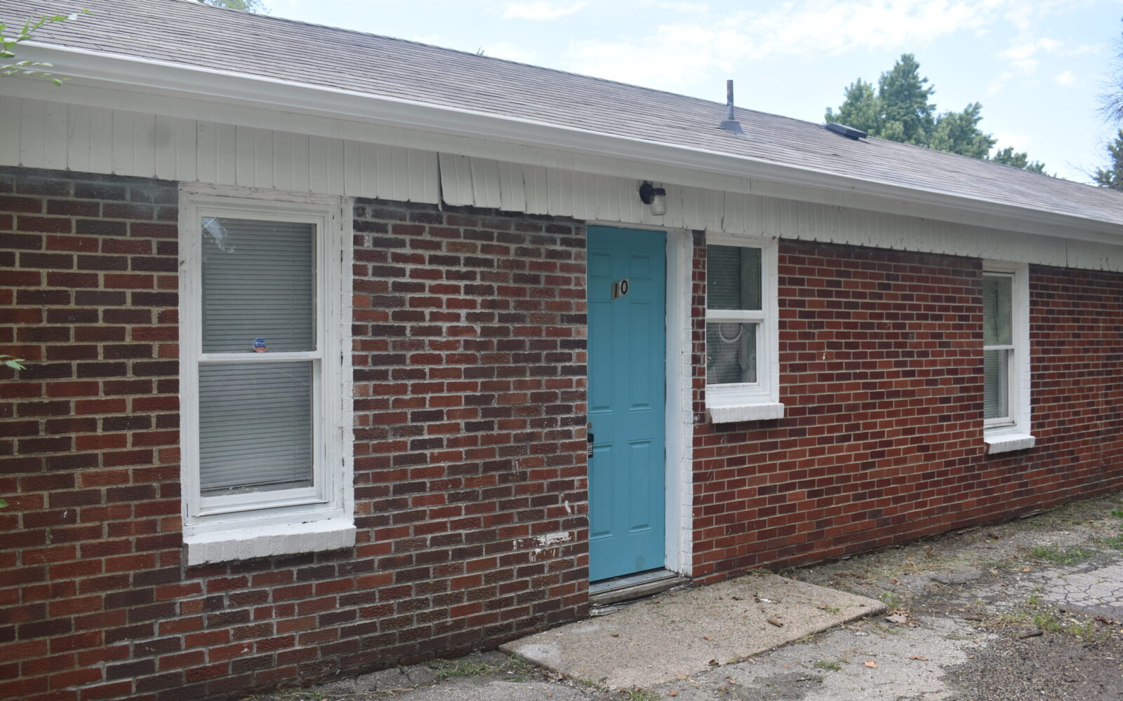 A red brick house with a light blue door numbered "10." The house has a white roof trim and two windows with white frames, one covered with blinds and the other with a curtain. The ground outside is paved with concrete. Trees are visible in the background.