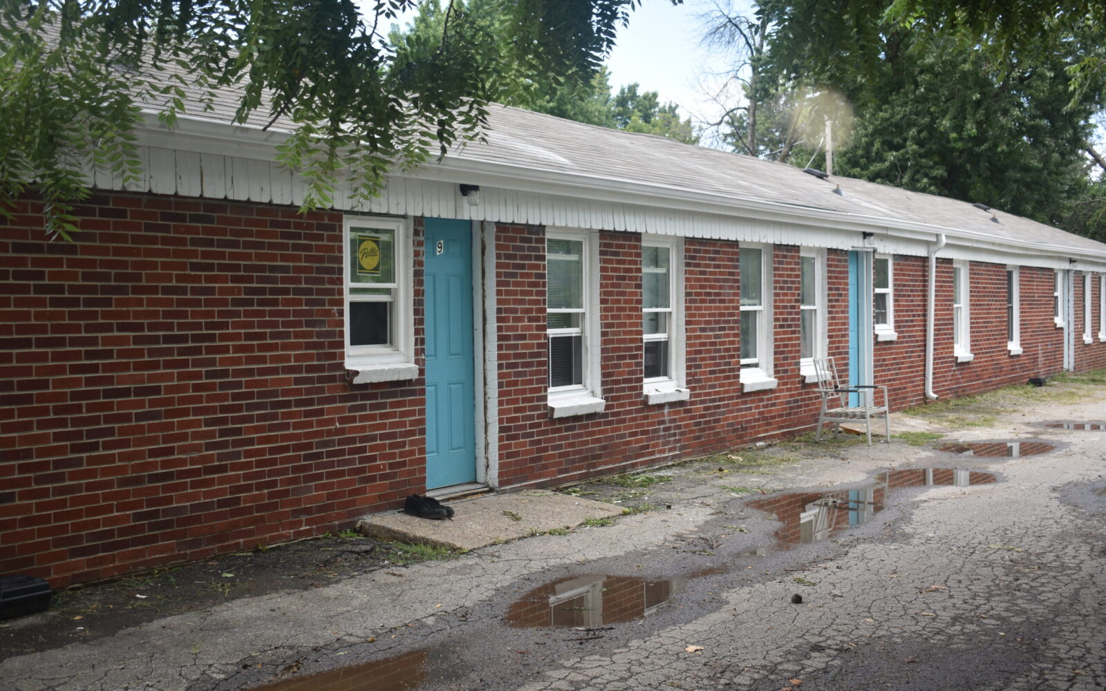 A single-story brick building with multiple turquoise doors and white-framed windows. The ground in front of the building is wet with puddles on the cracked pavement. Trees with green leaves hang overhead.