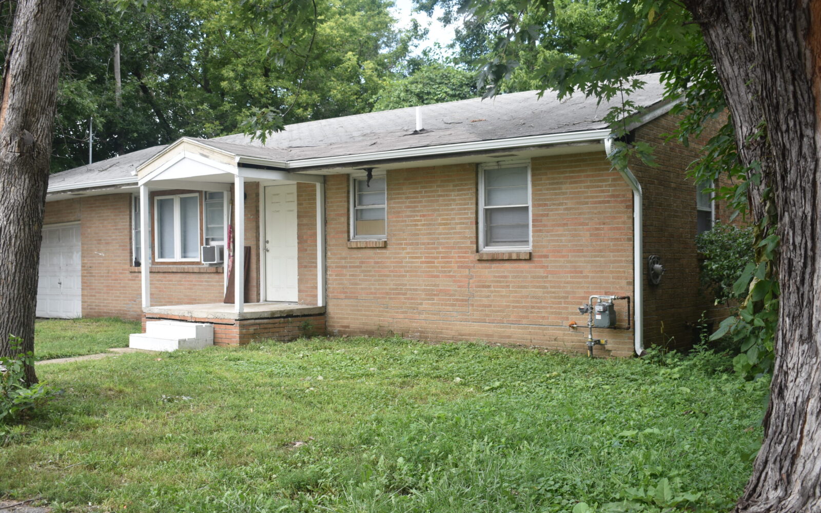 A single-story brick house with a white front door, small porch, and two windows at the front. The yard is grassy and partially overgrown. Two large trees frame the sides of the image. The house has an attached garage on the left side.