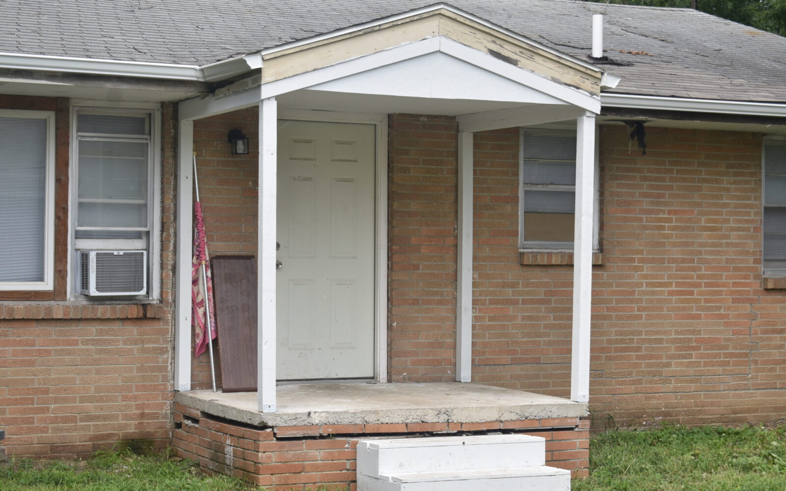 A small covered front porch with white pillars and steps, leading to a light-colored door on a brick house. Two windows with screens are visible on either side of the door, and an air conditioning unit is installed in the left window. There's a grassy lawn in front.