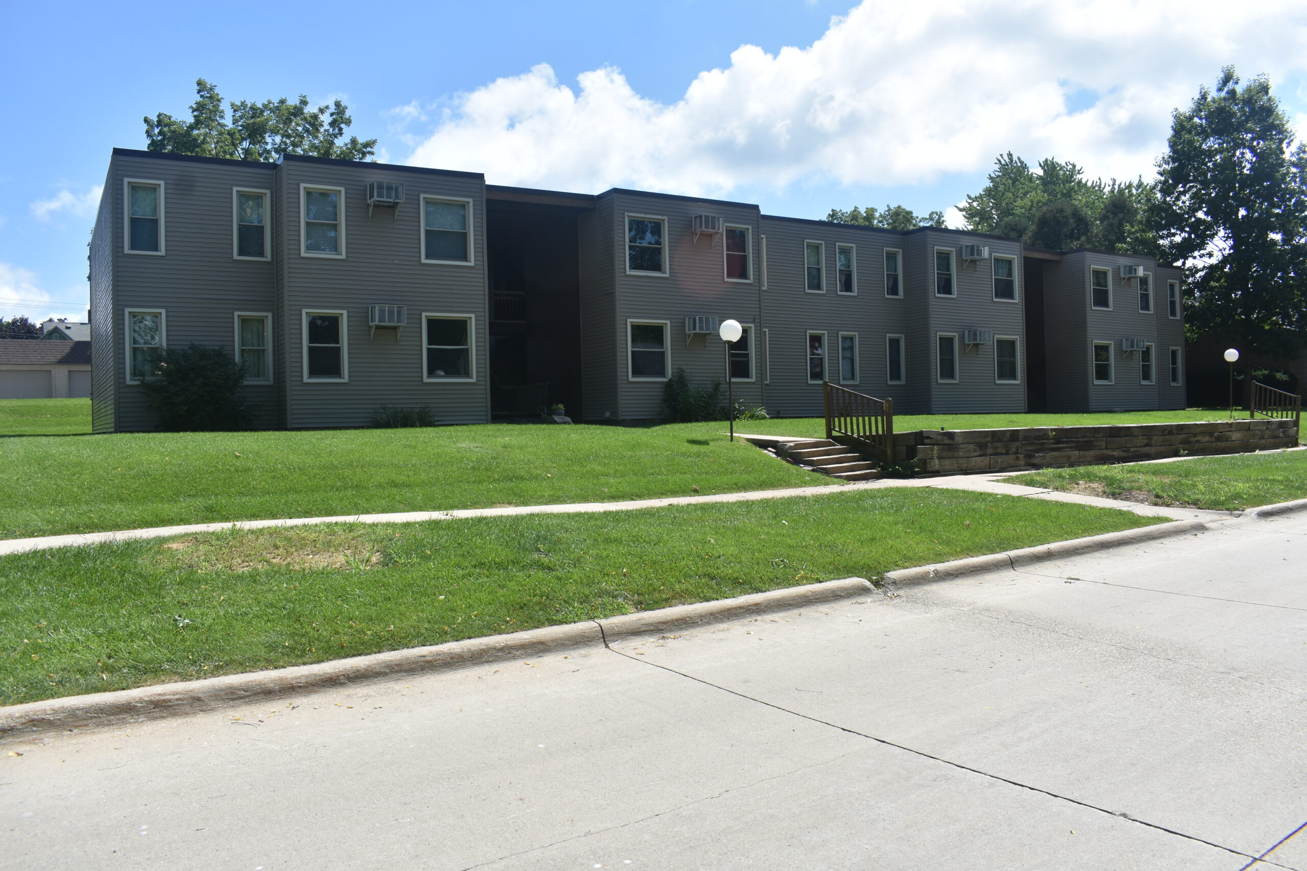 A two-story, U-shaped apartment building with gray siding and numerous windows sits behind a well-maintained, grassy lawn. A concrete pathway and steps lead from the sidewalk to the building's entrances. Trees and a partly cloudy sky are in the background.