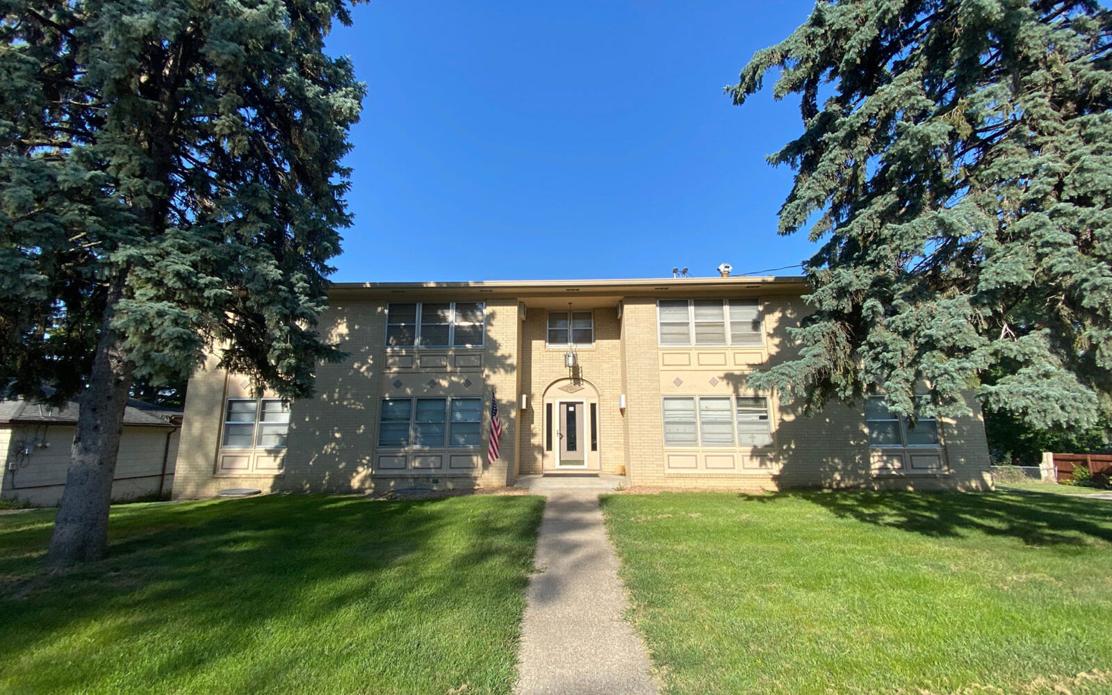 A two-story beige brick building with a central arched entrance and white front door. Tall evergreen trees flank the walkway leading to the entrance. The scene is well-lit with clear blue skies and a well-maintained green lawn. An American flag is displayed near the door.