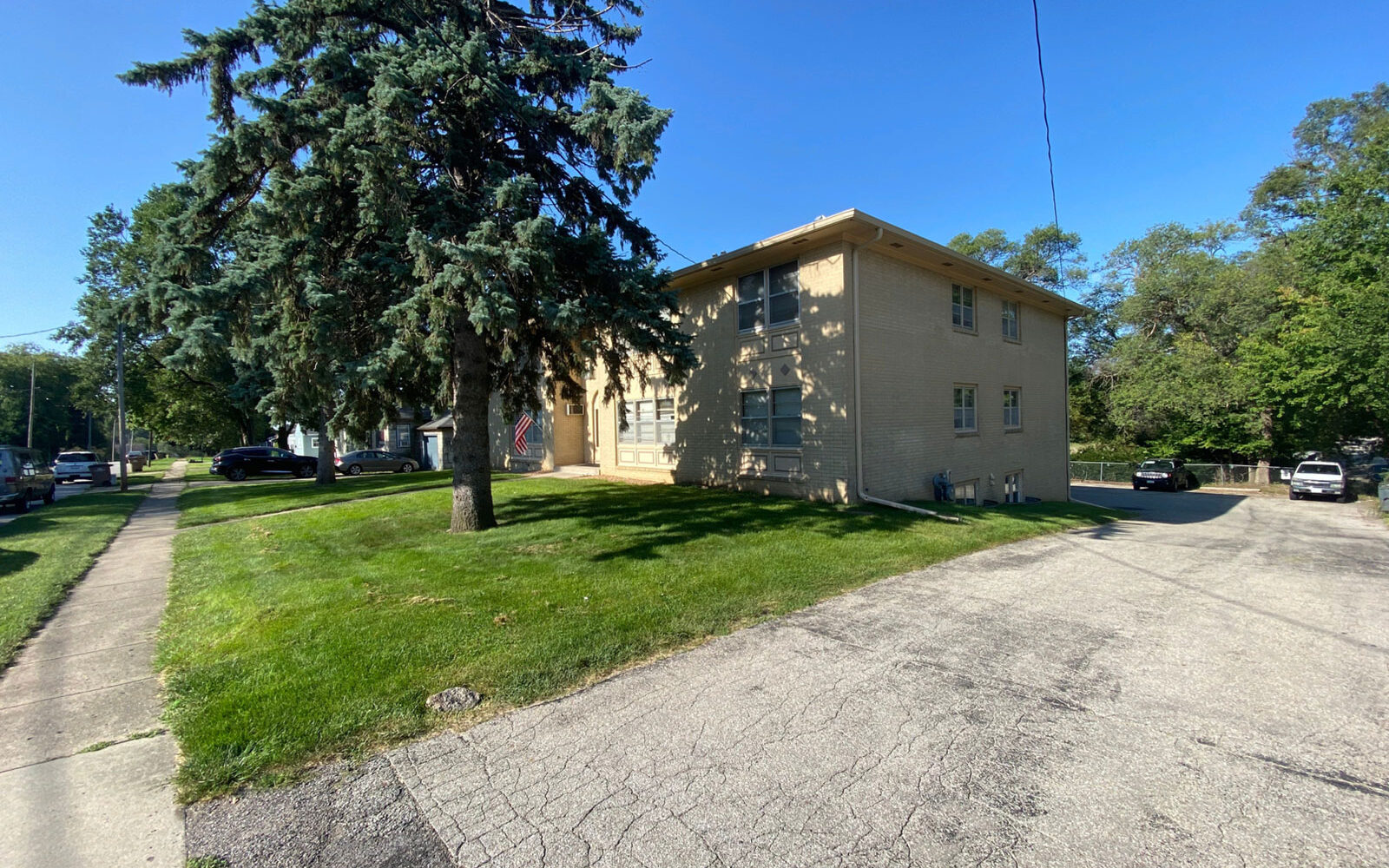 A two-story beige building is situated on a corner lot, surrounded by a lawn and paved driveway. A tall pine tree stands near the building, and several cars are parked in the driveway and along the street. The sky is clear and blue.