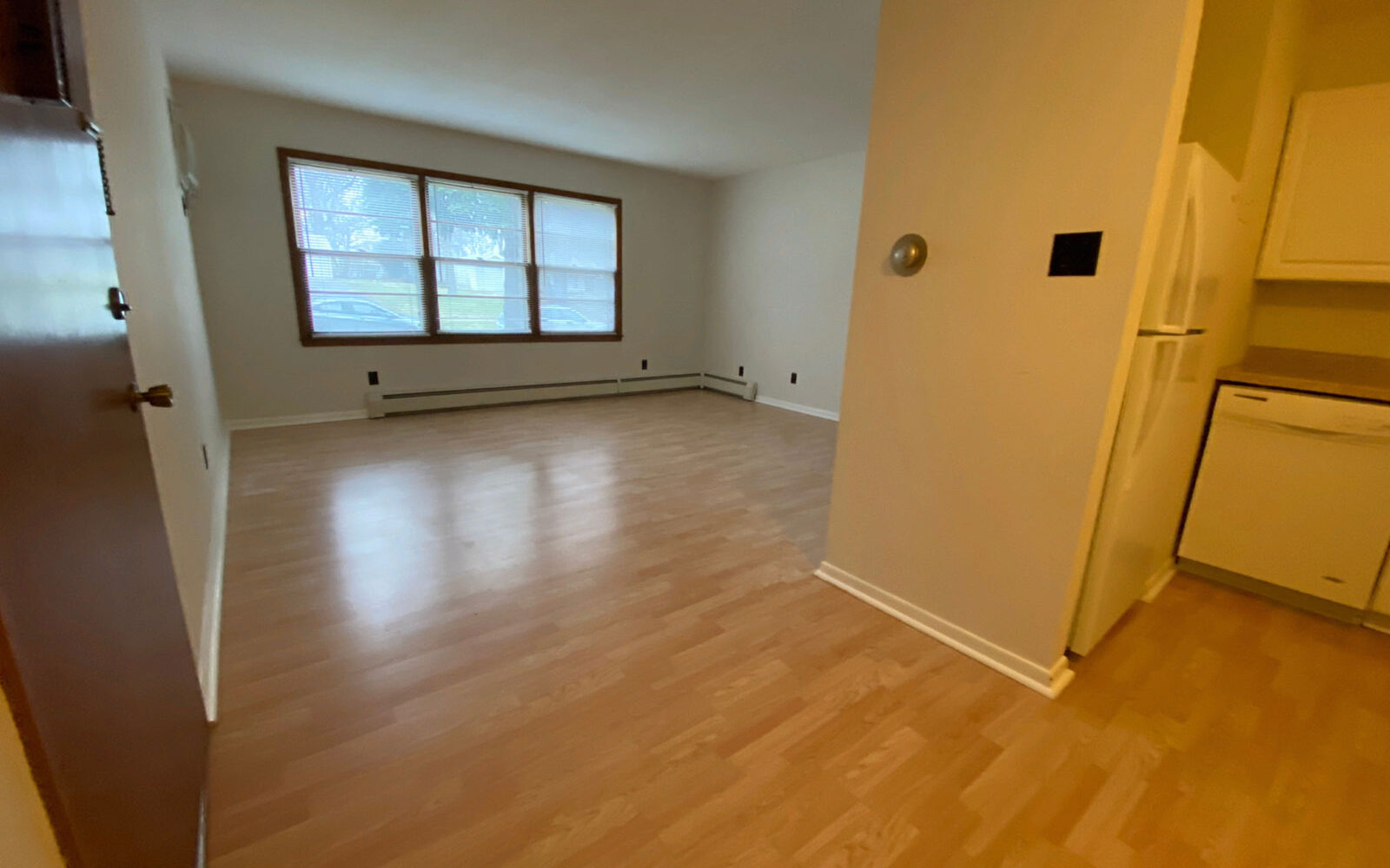 A vacant living room with light wooden flooring and cream-colored walls. Large windows line one wall, allowing natural light to fill the space. A partial view of a kitchen with white appliances is visible to the right.