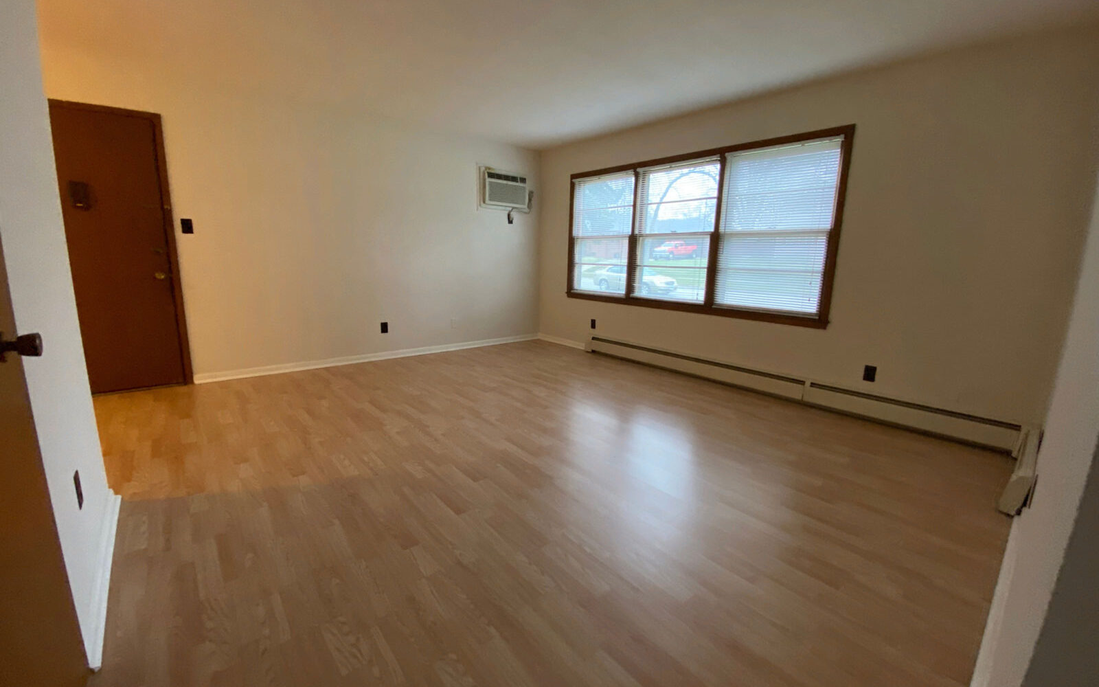 An empty room with light wood laminate flooring, a large window with blinds, and cream-colored walls. A door is situated on the left side, and a wall-mounted air-conditioning unit is positioned above a baseboard heater beneath the window.