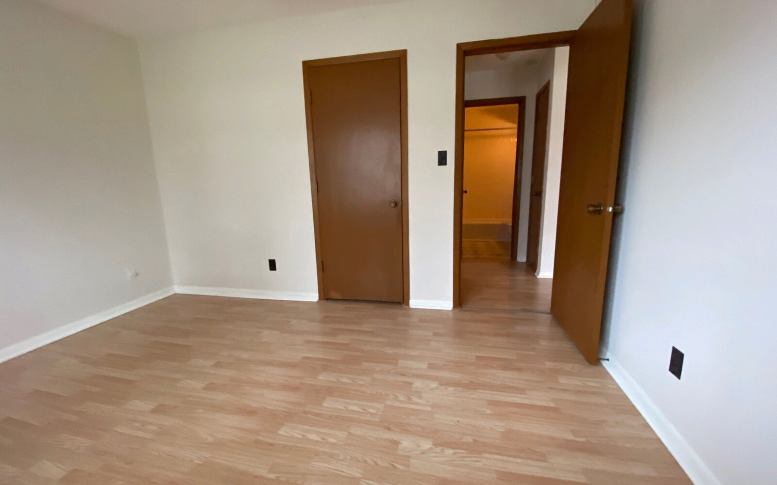 A small, empty room with light wooden flooring and white walls. Two wooden doors are visible: one closed and one open, leading to a hallway with a lit area. There is a ceiling fan partially visible at the top edge of the image.