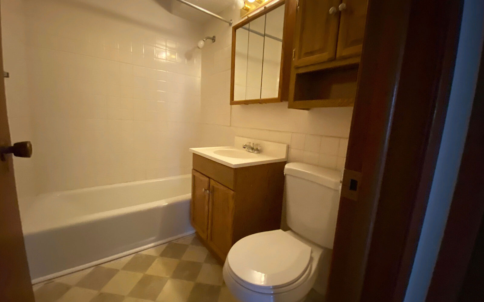 A bathroom with a bathtub-shower combination on the left, a vanity with a sink in the center, and a toilet on the right. Above the sink is a mirror with a wooden cabinet, and the floor has a checkered tile pattern. The room is illuminated by bulb lights.