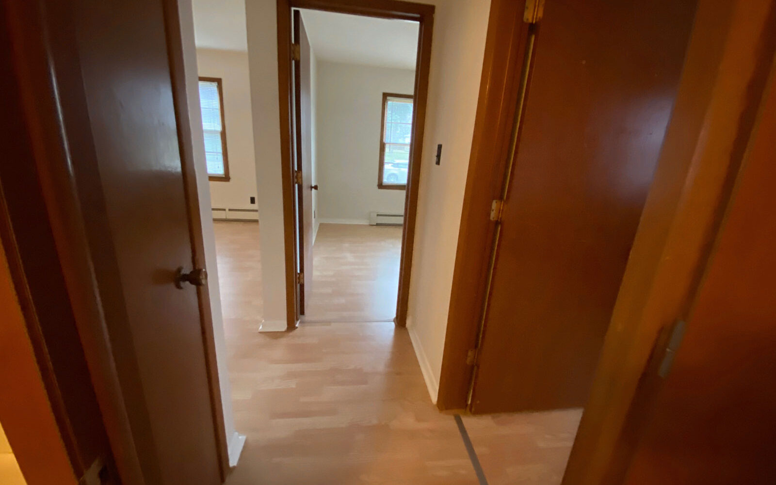 View of a hallway with light wood flooring, two open doorways leading to separate rooms with large windows, and a partially visible door on the right. The walls are painted light beige, and the space appears to receive ample natural light.