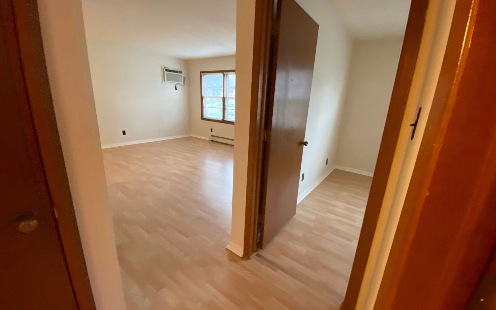 A view from a doorway into an empty apartment room with light-colored wooden flooring. There is a window and a wall-mounted air conditioner in the far wall, and a glimpse into another room on the right. The space appears bright and clean.