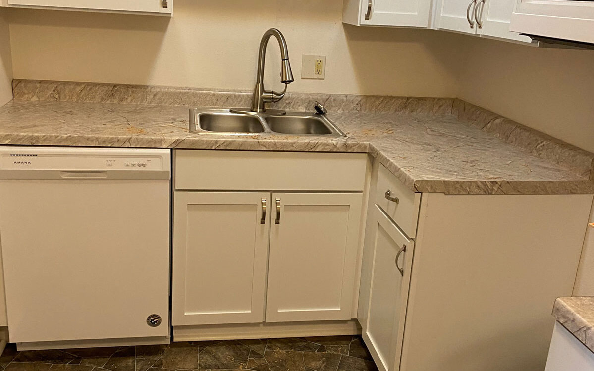 A small kitchen features white cabinetry, a dual-basin stainless steel sink with a chrome faucet, a dishwasher, and a beige countertop that extends into a corner. The floor is covered with patterned tiles in shades of brown and gray.