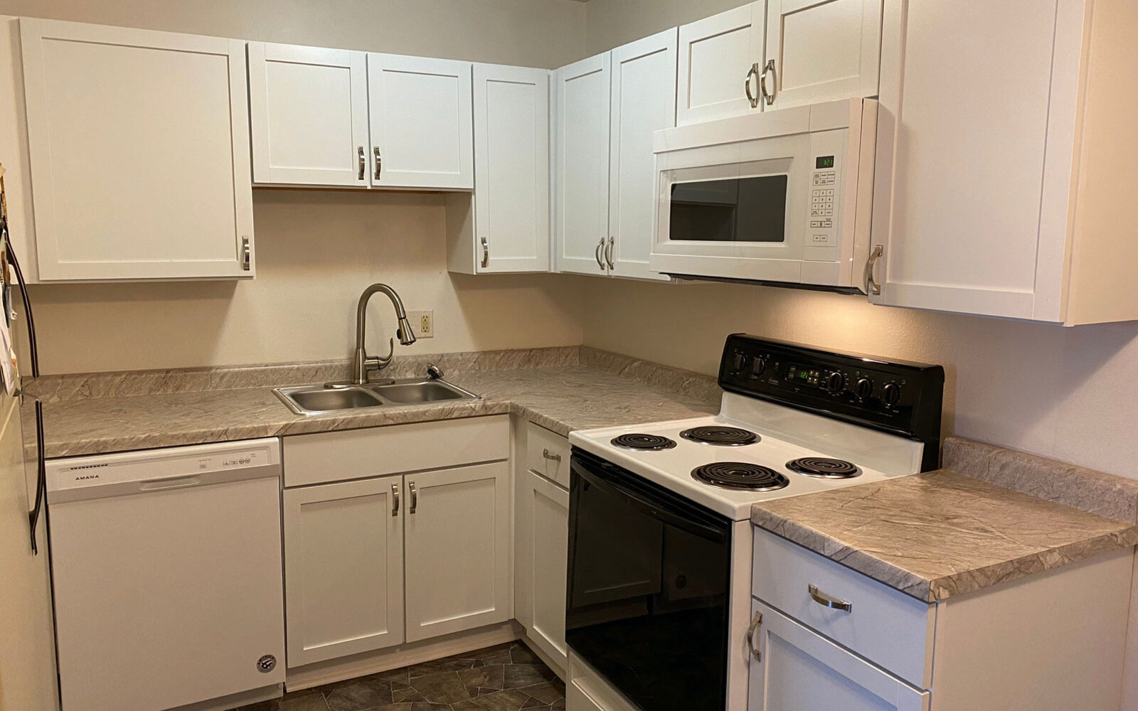 A modern kitchen with white cabinetry, granite countertops, and stainless steel sink. Includes a white dishwasher, a white microwave, and a black electric stove. The floor is covered with dark patterned tiles.