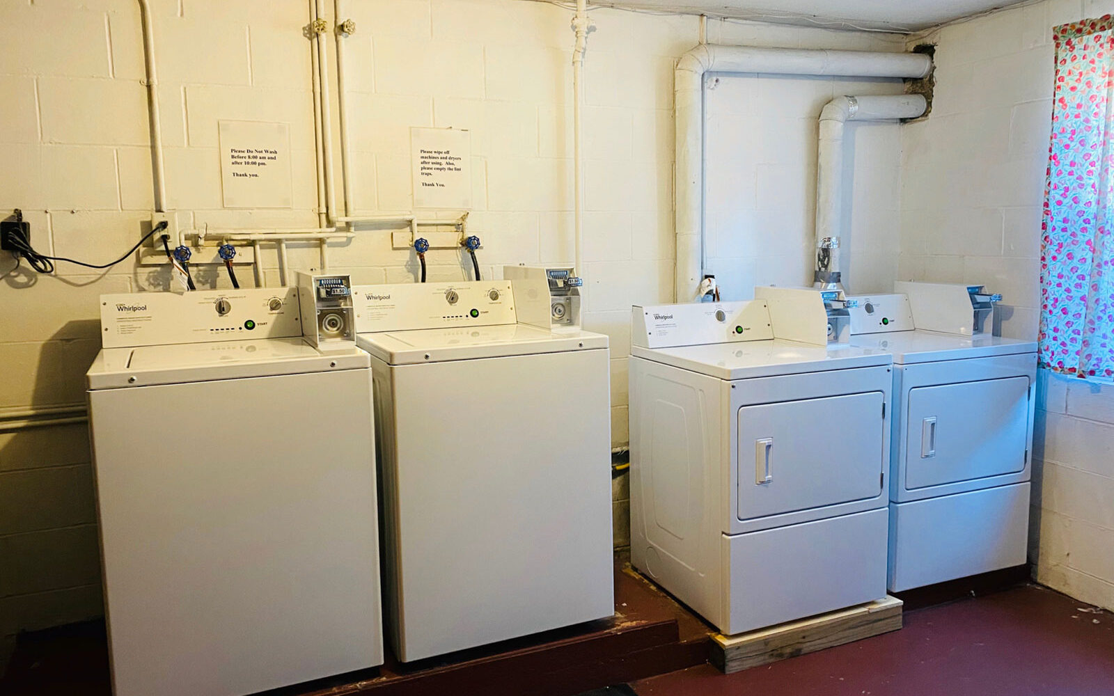 A bright laundry room with white brick walls features two washing machines and two dryers. Each set is topped with folded instructions or notices. The floor is red-brown, and a floral curtain partially covers a nearby window. Pipes run along the walls and ceiling.