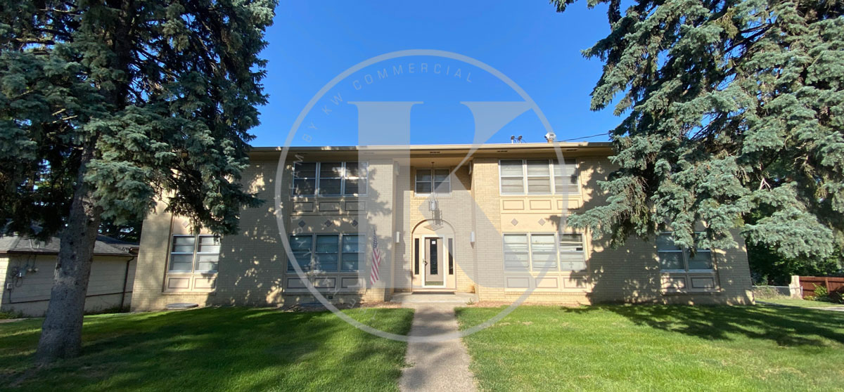 A two-story beige building with a central entrance and a large front yard is flanked by tall trees. The building features multiple windows and has a concrete pathway leading to the front door. The sky is clear and blue, suggesting a sunny day.