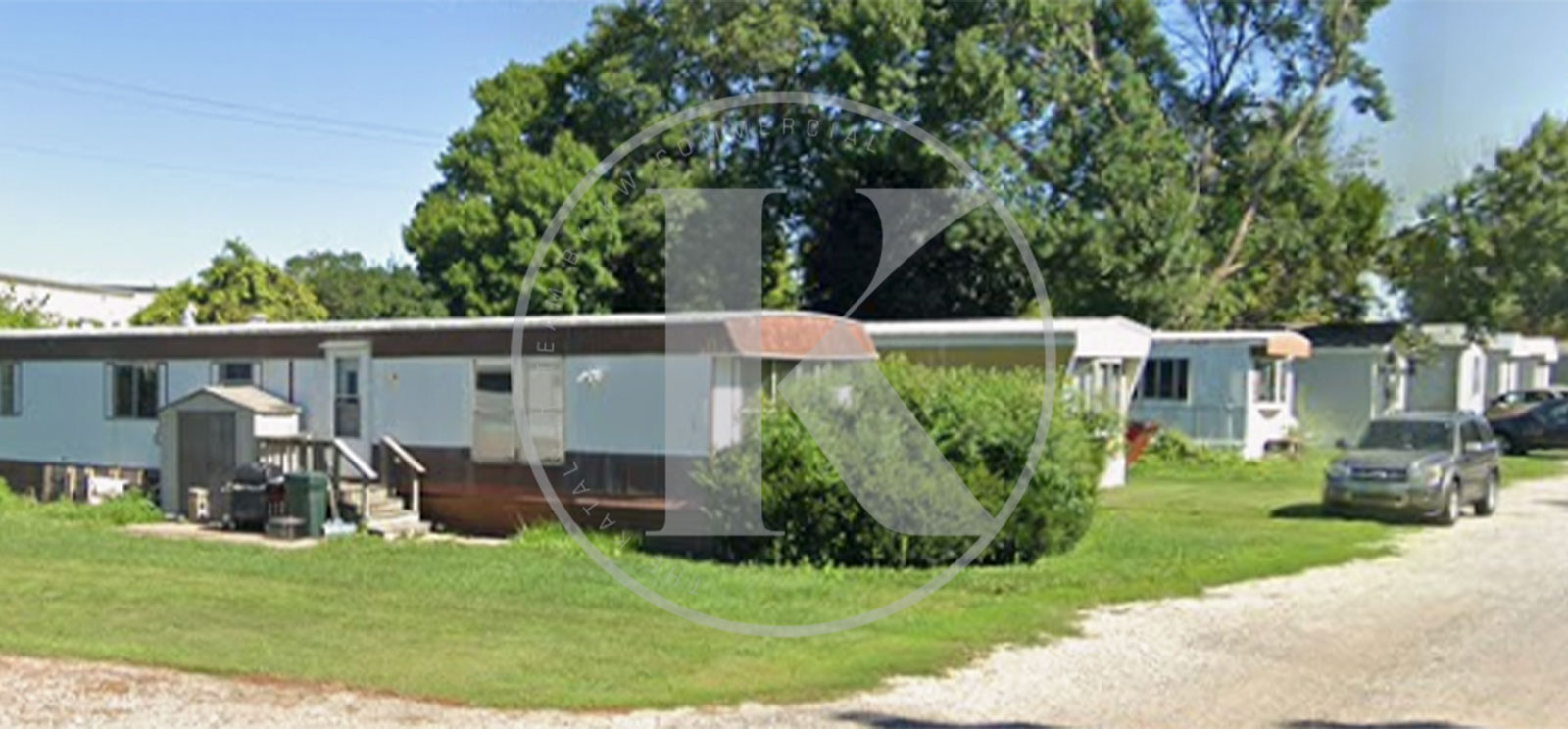 A row of mobile homes with white exteriors sits along a gravel path, surrounded by lush green trees and grass, under a clear sky. A car is parked near the homes.