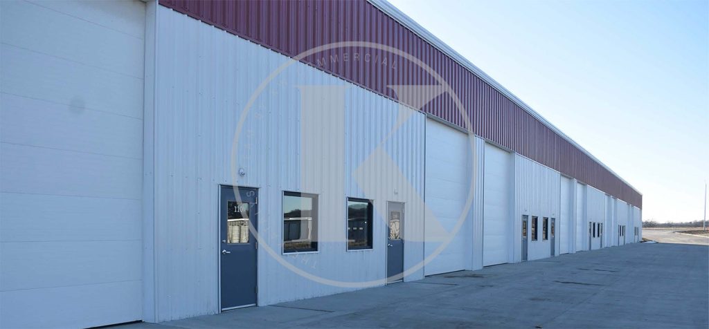 Long white industrial building with a red roof and multiple doors and windows. The structure is set against a clear blue sky along a paved surface.