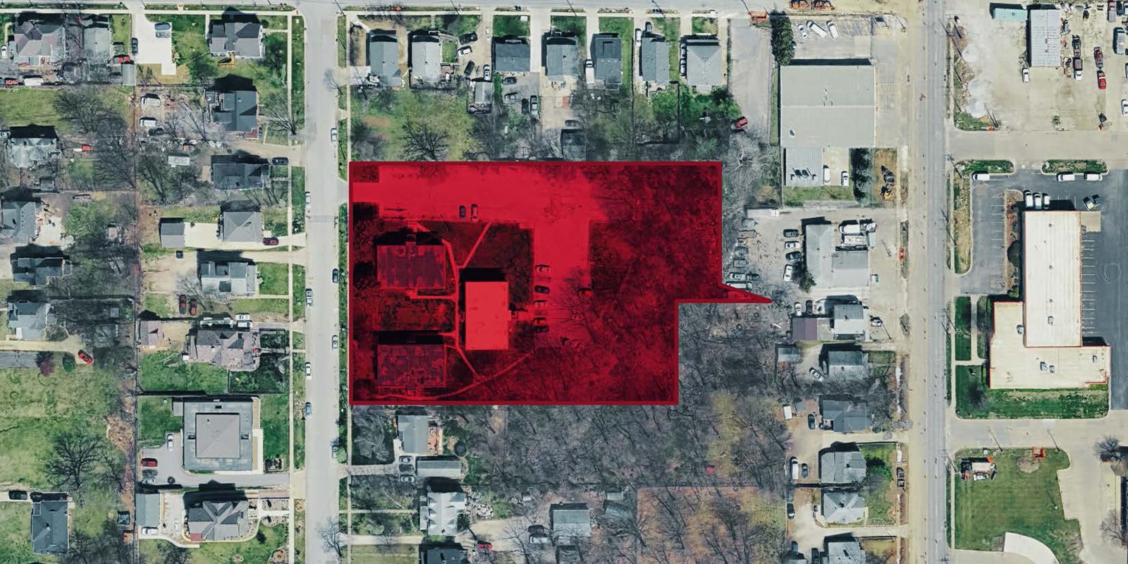 Aerial view of a suburban neighborhood with a red-highlighted area encompassing several residential properties, trees, and a road intersection. Surrounding streets and houses are visible.