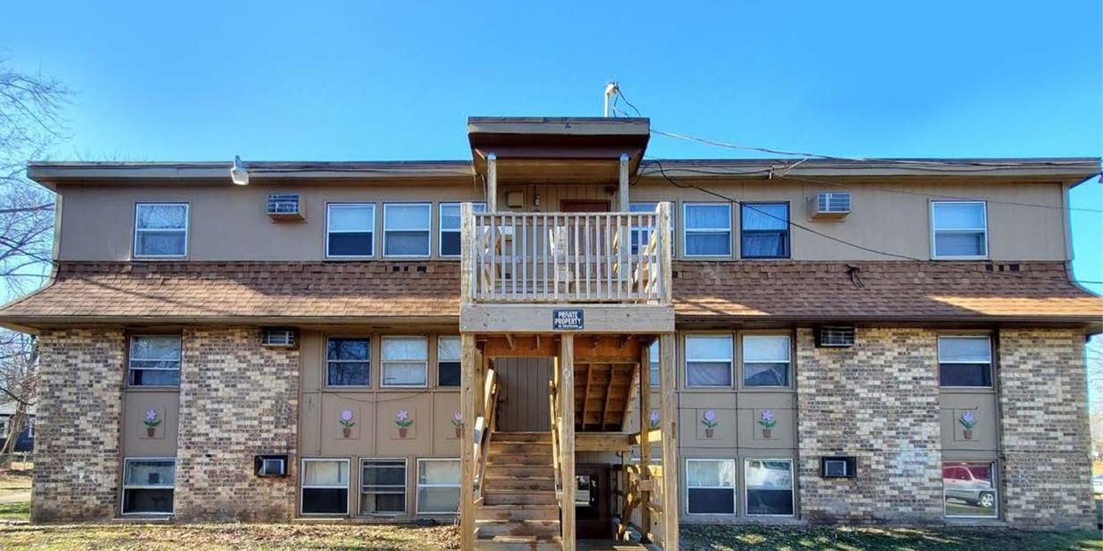 Two-story apartment building with brick and siding exterior. Central wooden staircase leads to the second floor. Windows line both levels. Air conditioning units visible under windows. A sign is on the railing of the upper balcony. Clear blue sky.