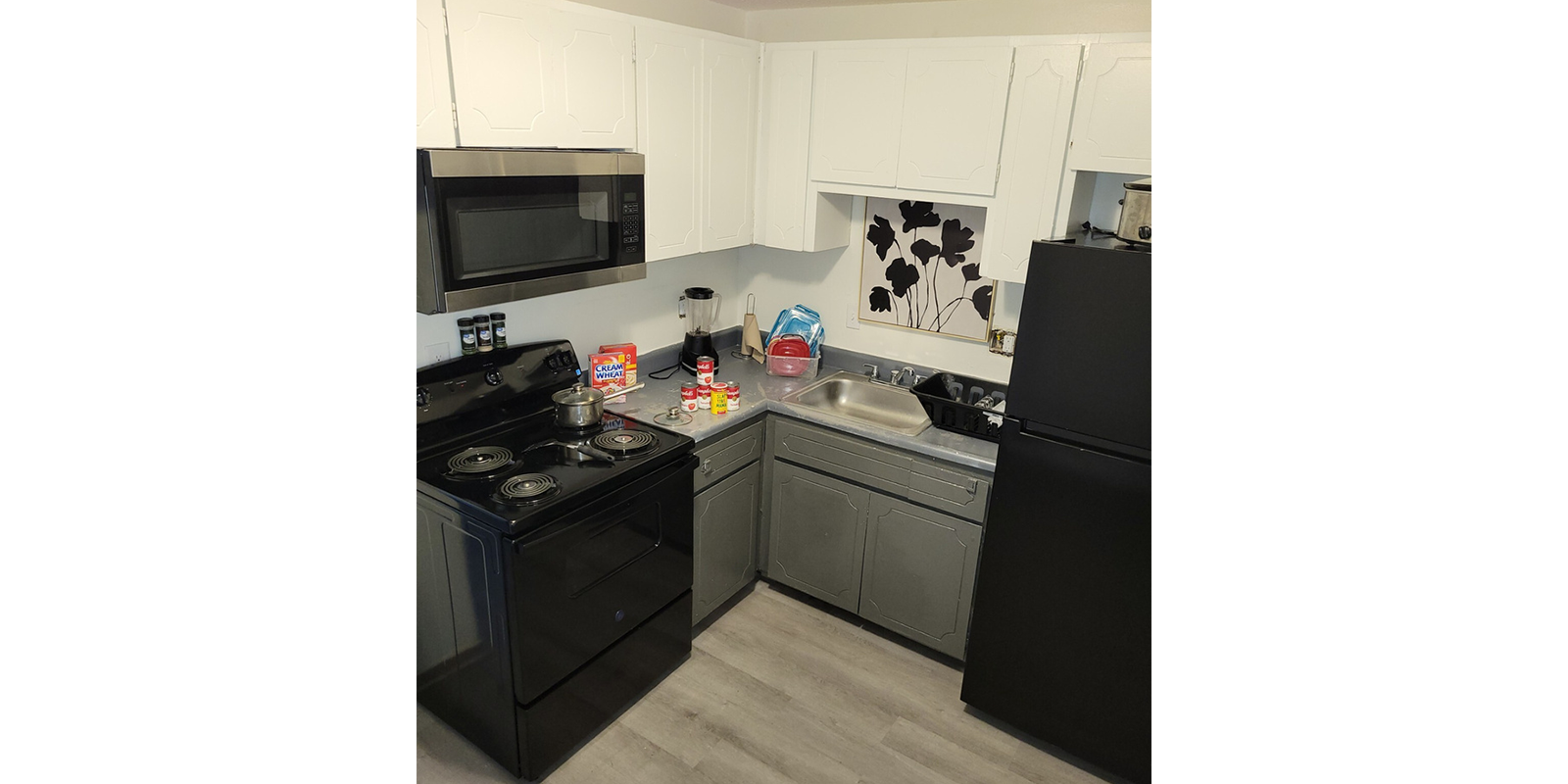 A compact kitchen features a black stove with pots, a black microwave above, and a black refrigerator. The counter holds food items, a blue basket, and cleaning supplies. The sink is stainless steel, and cabinets are white. The floor is light-colored laminate.