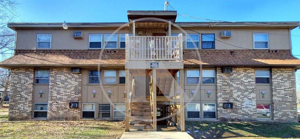 Two-story residential building with a brown roof and brick facade. The entrance features a central staircase leading to a wooden balcony. Windows line the upper and lower levels, with small decorative plants below some windows. A grassy lawn surrounds it.