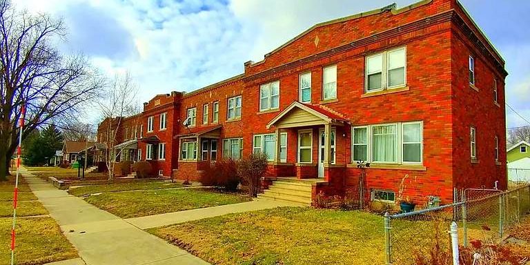 A row of two-story, red-brick apartment buildings with white-framed windows and small front porches sits along a sidewalk under a partly cloudy sky. Some leafless trees and lawns are visible in front of the buildings.