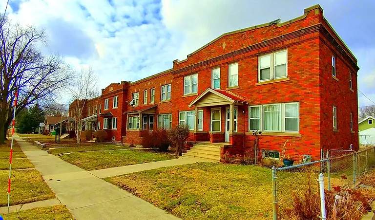 A row of two-story, red-brick apartment buildings with white-framed windows and small front porches sits along a sidewalk under a partly cloudy sky. Some leafless trees and lawns are visible in front of the buildings.