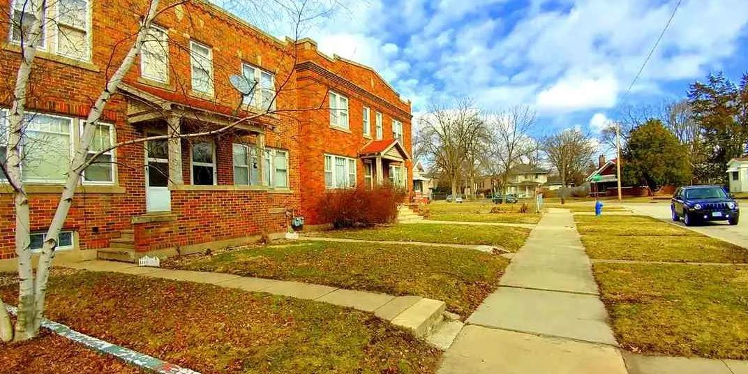 A row of red-brick townhouses on a suburban street with patches of grass, leafless trees, a blue car parked on the right, and partly cloudy skies overhead.