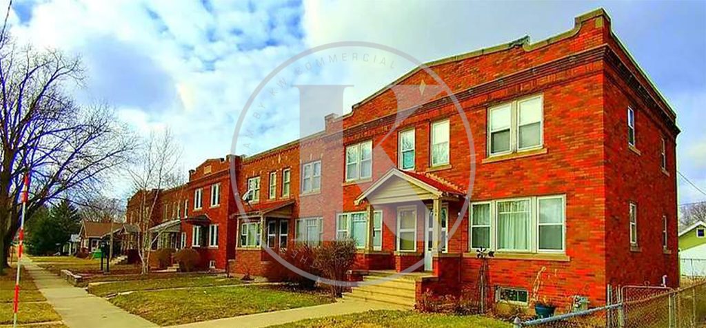 A row of red brick apartment buildings with white-trimmed windows, a small set of stairs leading to the entrance, and a sidewalk bordered by grass under a partly cloudy sky.