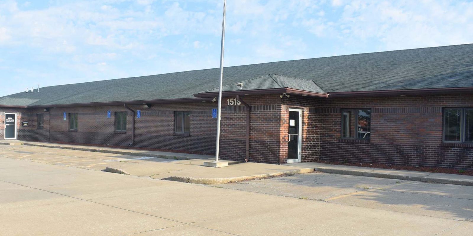 A single-story brick building with a sloped roof, an American flag on a tall pole in front, and an empty parking lot under a partly cloudy sky. The building's address, 1515, is visible above the entrance.