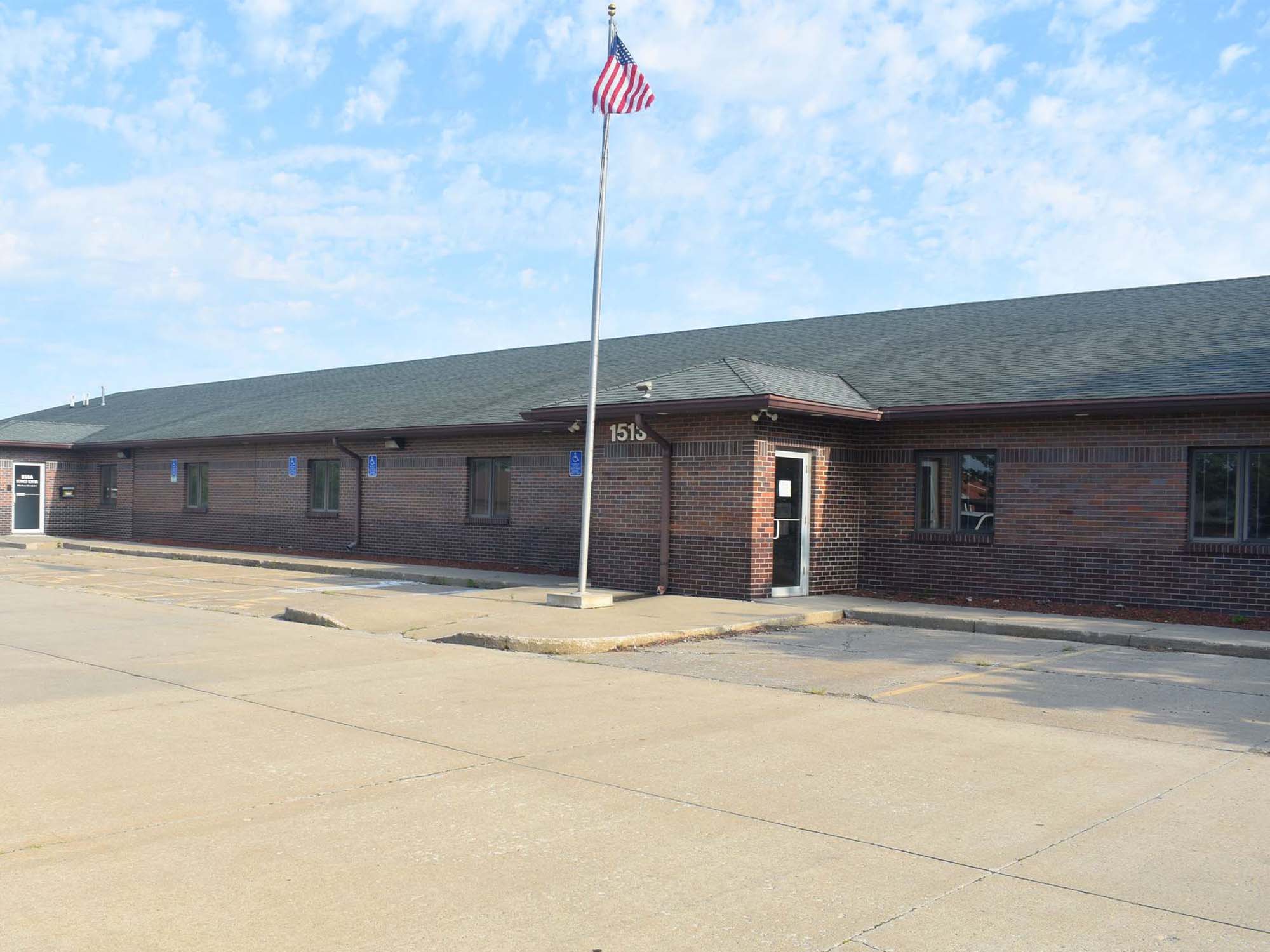 A single-story brick building with a sloped roof, an American flag on a tall pole in front, and an empty parking lot under a partly cloudy sky. The building's address, 1515, is visible above the entrance.