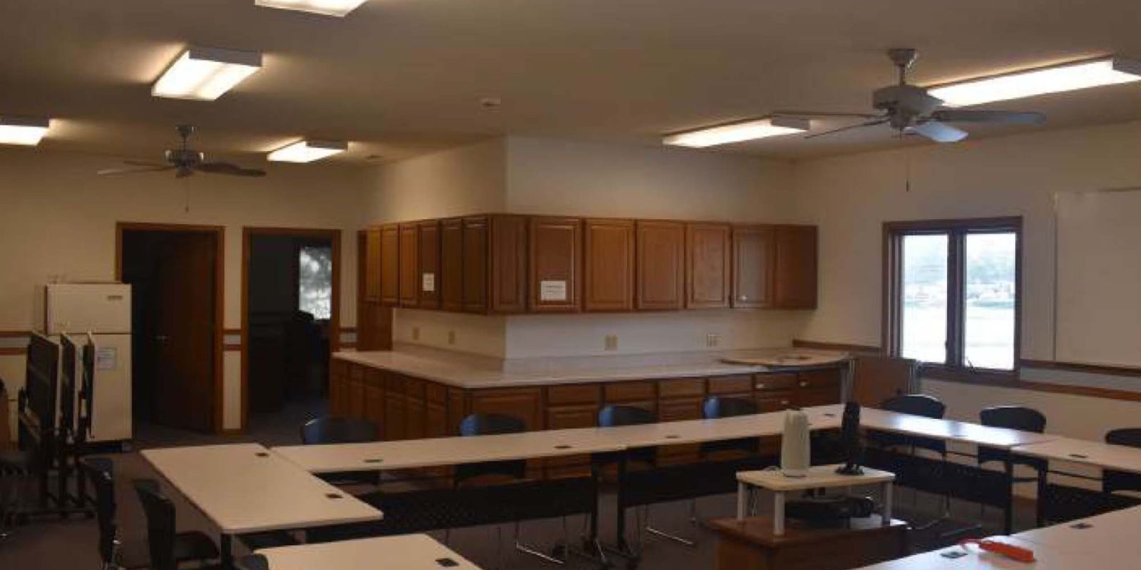 A classroom with U-shaped tables, blue chairs, a large whiteboard, cabinets along the back wall, windows, and fluorescent ceiling lights. A bottle of sanitizer sits on one of the tables.