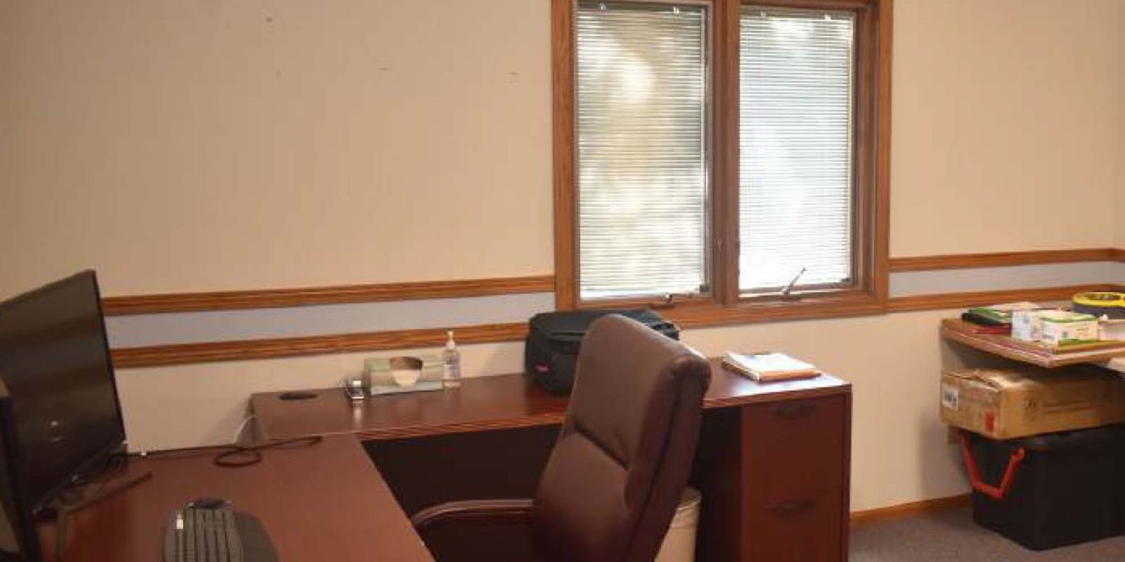 An office with a brown desk, office chair, computer monitor, keyboard, and various items. There’s a window with blinds, a filing cabinet, and boxes stacked in the corner of the carpeted room.