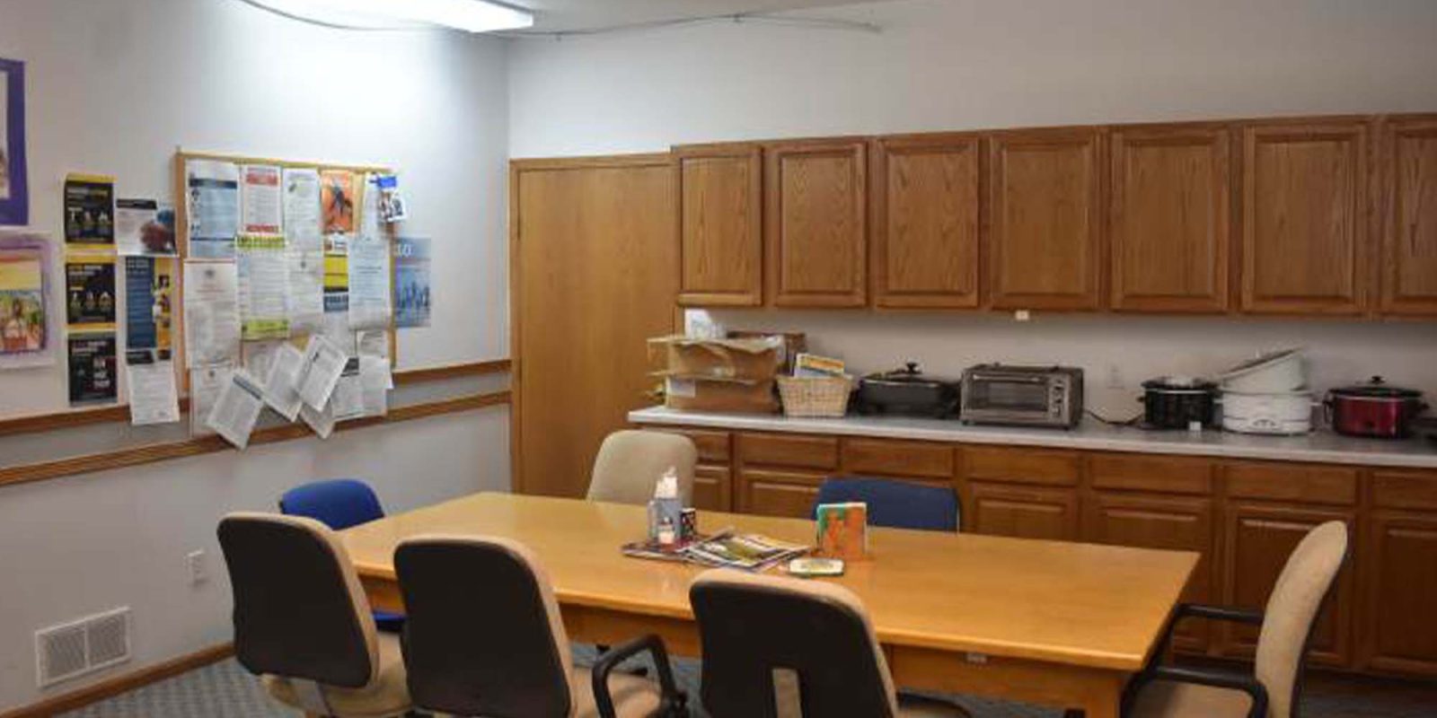 A meeting room with a wooden table surrounded by six chairs, a wall of cabinets, kitchen appliances including slow cookers, and a bulletin board covered with papers and notices.