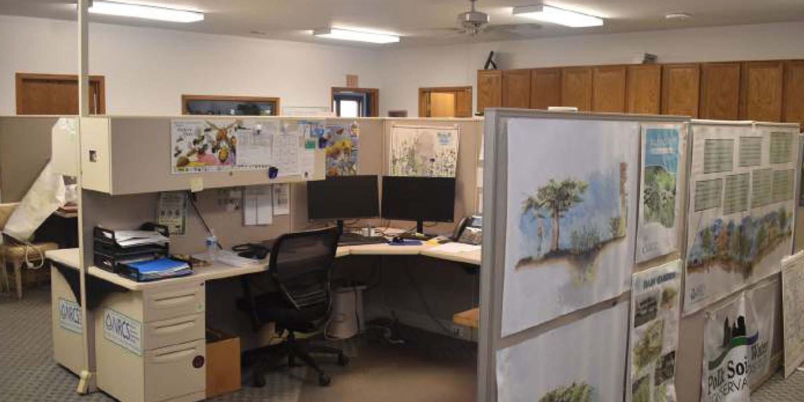 An office cubicle with two desks, computer monitors, paperwork, and office supplies. Large illustrated plans and posters are displayed on one side of the cubicle. The space is well-lit with overhead lights.