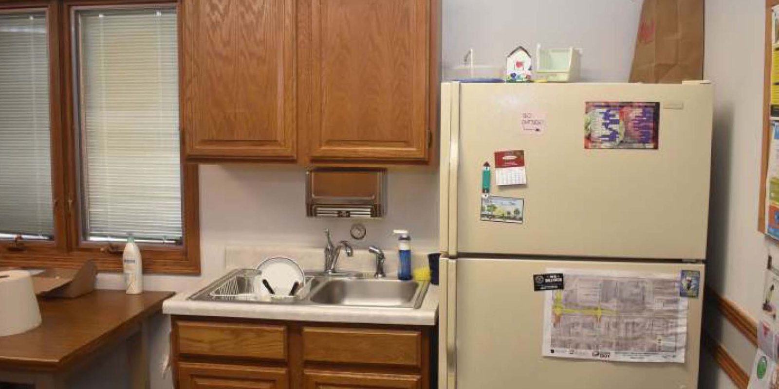A small kitchen area with a sink, wooden cabinets, a beige refrigerator with magnets and papers, a window with blinds, and a paper bag on top of the fridge. A table with paper towels is on the left.