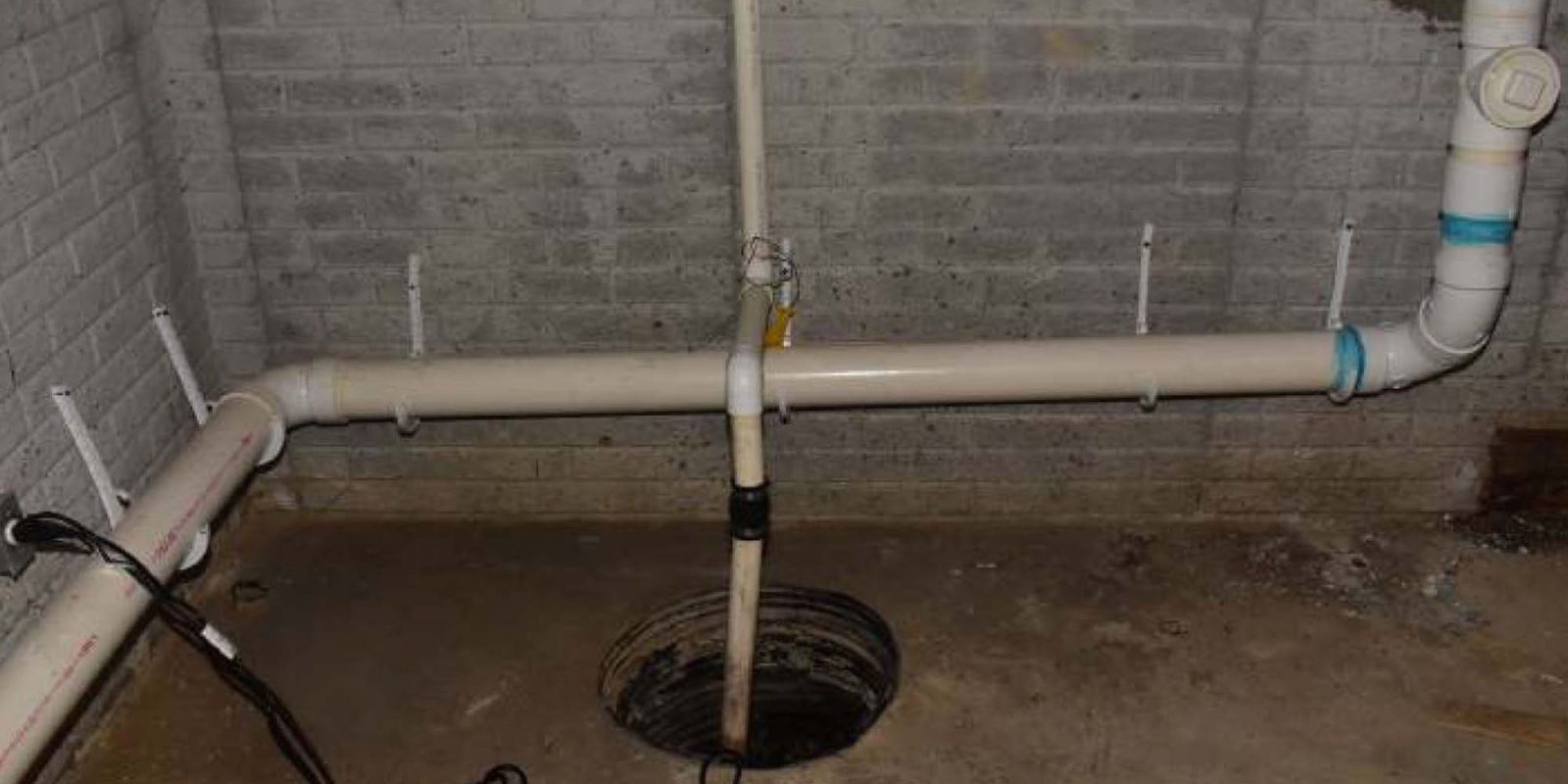 A basement with exposed brick walls, a concrete floor, visible white PVC pipes, and an open sump pit in the center. Electrical cords and some debris are scattered on the floor.