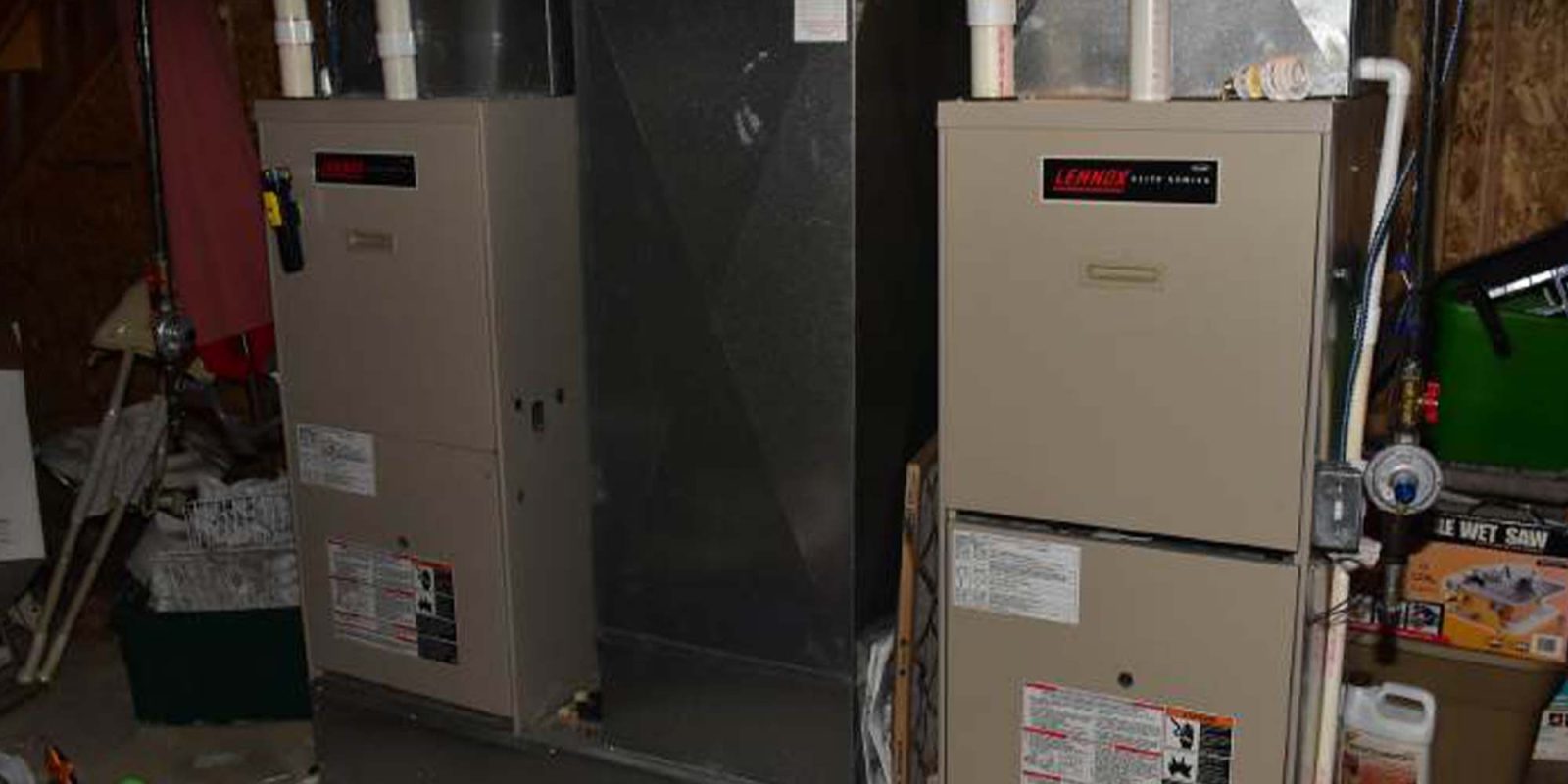 Two beige Lennox gas furnaces with white PVC pipes are installed side by side in a basement with concrete floors and exposed walls. Toys and storage boxes are visible nearby.